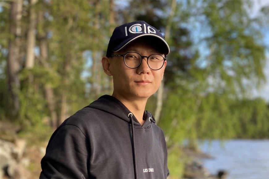 Man looking at camera with a cap hat on. Trees and water on the background. 