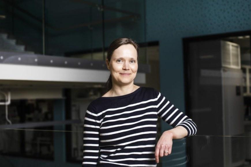 Half-length portrait of Minna Hankaniemi wearing a black and white striped shirt. There is a turquoise wall, large windows and glass railings in the backround.