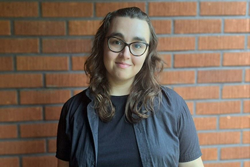 Magdalena Celuch dressed in a dark shirt with a brick wall in the background.Magdalena Celuch pukeutuneena tummaan paitaan, taustalla tiiliseinä.