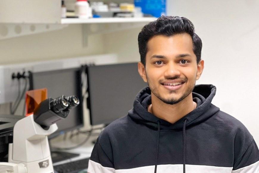 A person in a hoodie sits in a laboratory, smiling and looking at the camera.