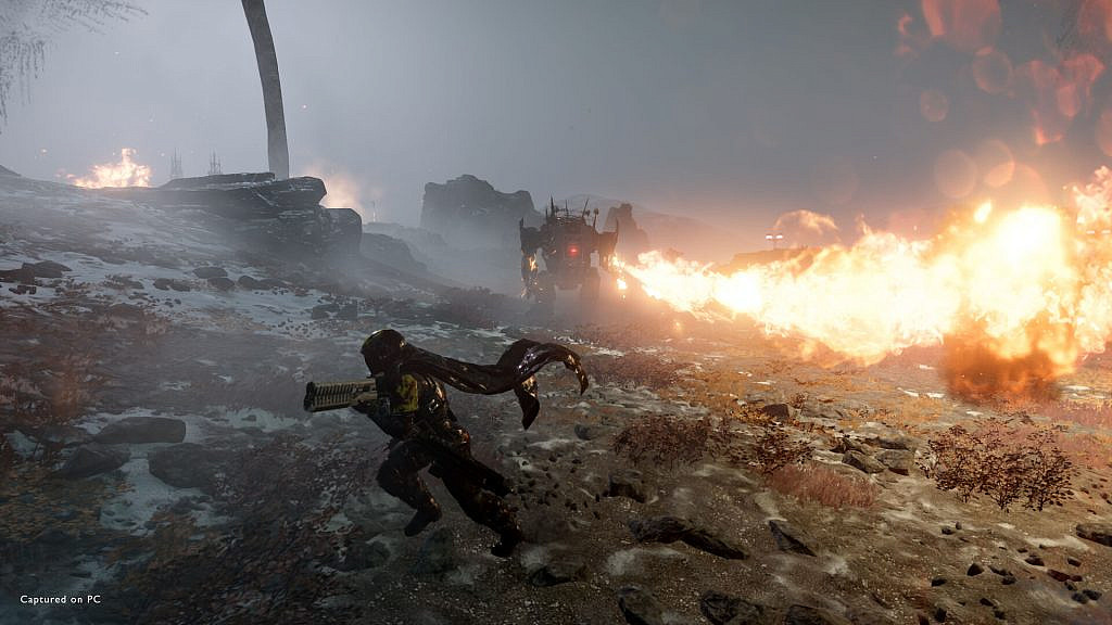 A Helldiver soldier is running away from a huge robot enemy/Automaton in a rocky environment. The enemy is firing a flamethrower towards the soldier.