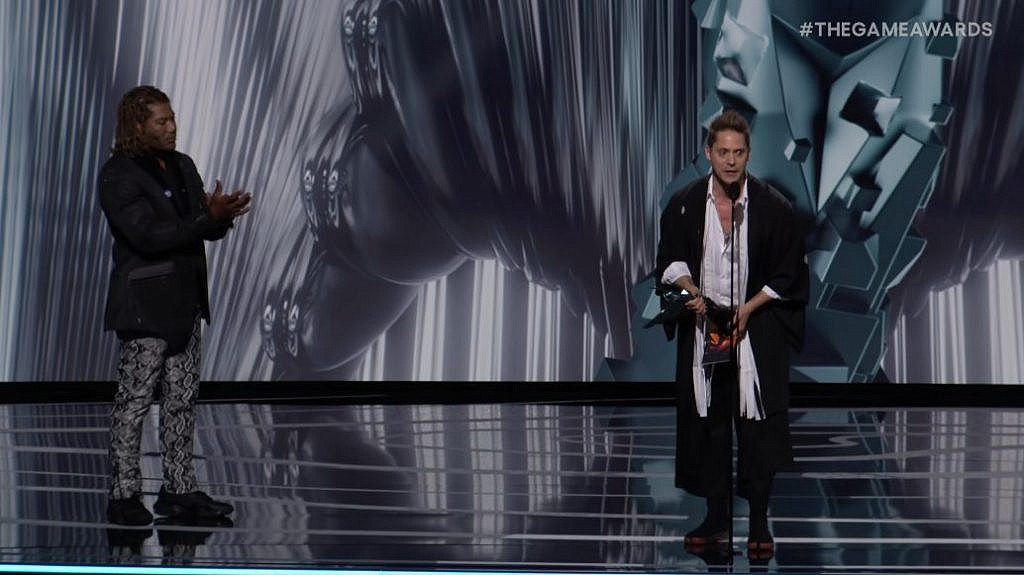 Two men on stage. Lef one is clapping and the right one is giving an acceptance speech with an award in hand