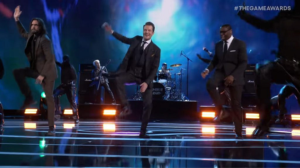 Three men in suits performing a coreographed dance onstage with a band playing in the background